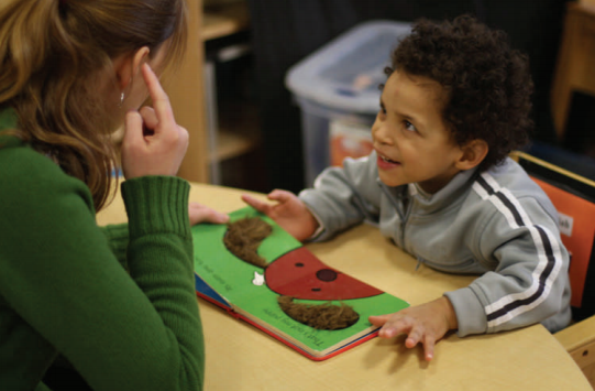 A boy and adult woman are sitting at a table. The boy has a children's book open before him. He is looking at the woman, smiling, and she is pointing to her ear.