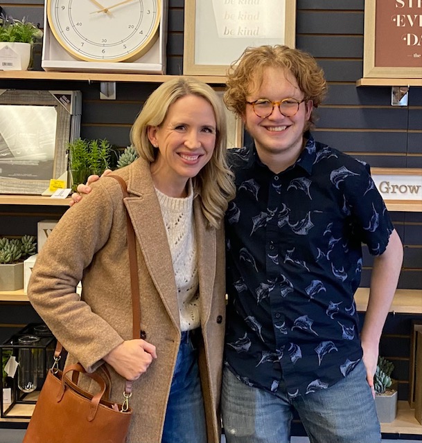 A woman and young man stand side by side, smiling. He has his arm on her shoulder.