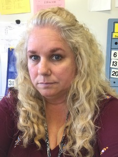 Head and shoulders of a woman with blonde hair, in front of a bulletin board.