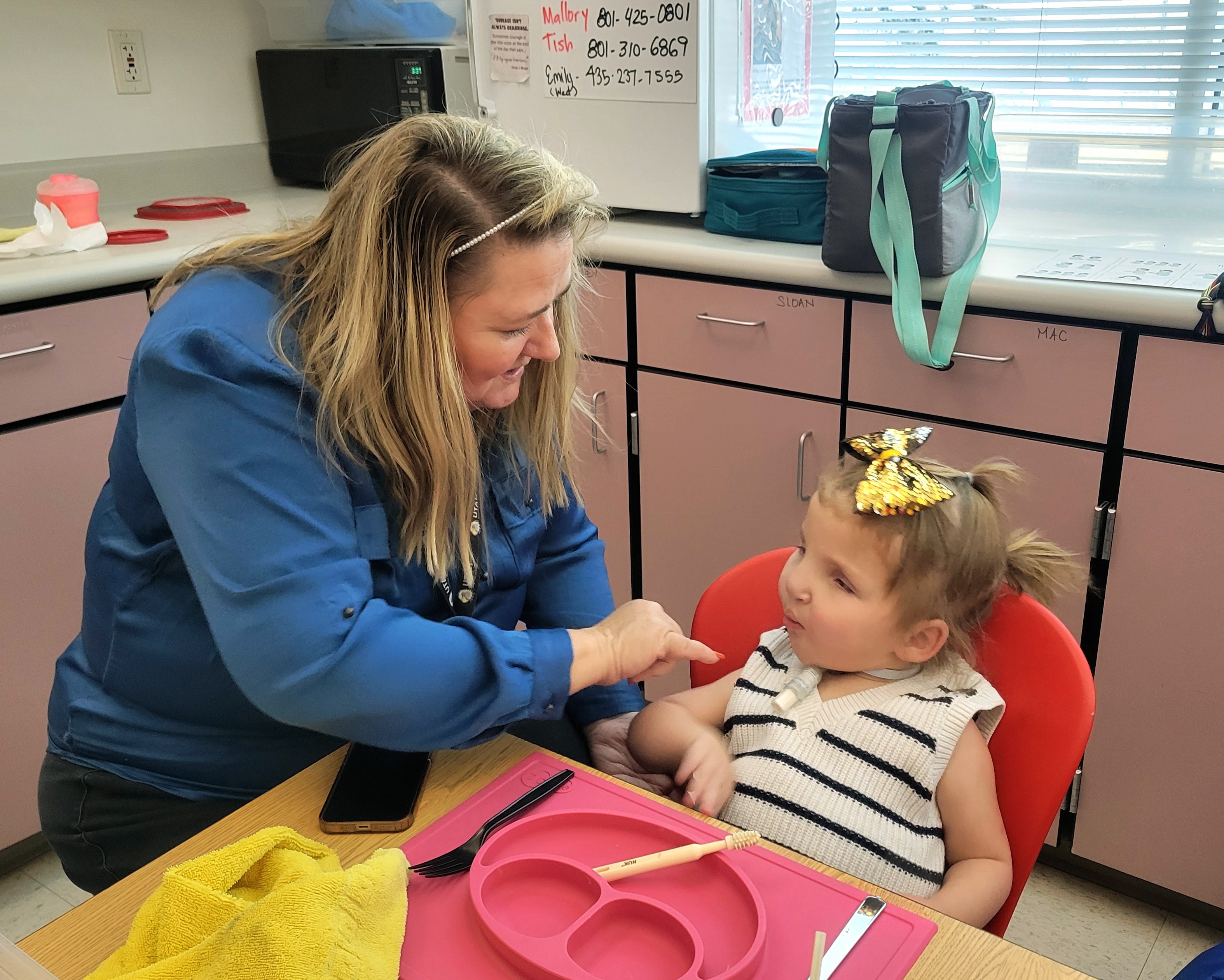 A young girl who is deafblind sits in a highchair. She has a tracheostomy tube in her neck. In front of her is an empty child’s plate and utensils. An adult female sits beside her. The adult faces the girl and extends her hand towards her.