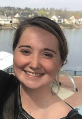 Close up photo of a smiling woman. In the background is a body of water with houses and trees on the other side.