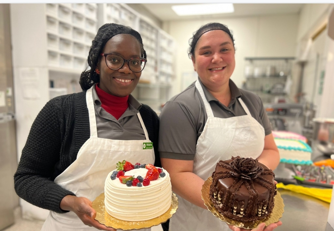 Two women standing next to each other wearing aprons and holding decorated cakes.