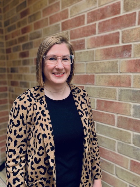 Smiling woman with blond hair and glasses, standing in front of a brick wall.